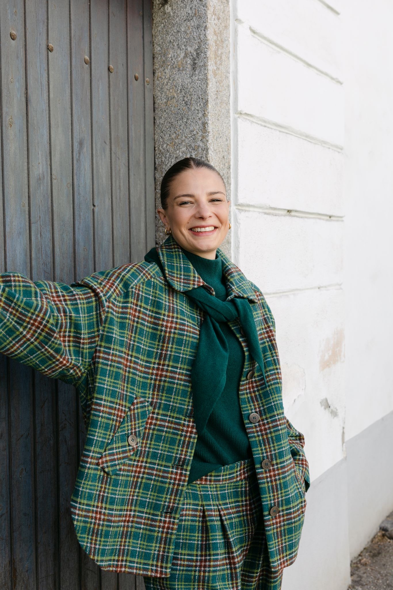 Grünes Karo-Set mit Rollkragenpullover und Schal, lachend an einer alten Holztür bei ZWEI auf der Spittelwiese in Linz